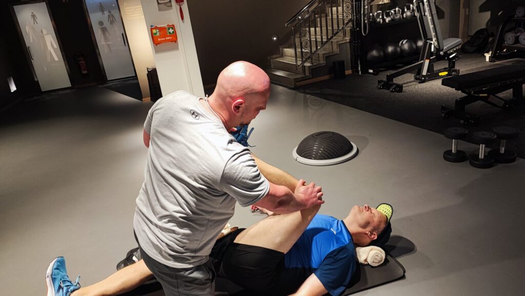 Ein professioneller Personal Trainer führt bei einem Kunden auf einer Trainingsmatte eine geführte Dehnübung zur Schmerzlinderung und Regeneration in Hamburg durch.